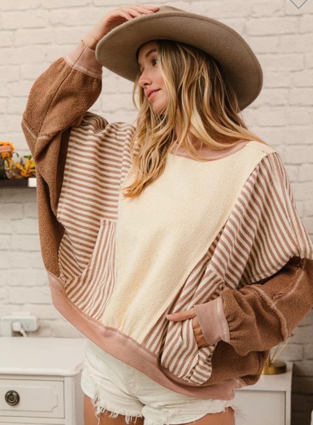 A female model wearing a beige and brown striped pullover with a relaxed fit, paired with white shorts and a wide-brimmed hat.