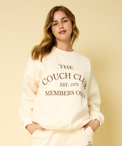 Model wearing a cream-colored sweatshirt with 'THE COUCH CLUB' and 'EST. 1971' printed in brown on the front, paired with matching pants.